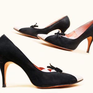 May include: A pair of black suede heels with a pointed toe and a small bow detail. The toe cap is a light color, and the heels are a slender, high stiletto style. The interior lining is a light pink color.