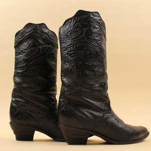 May include: A pair of black leather cowboy boots with intricate embossed patterns. The boots have a pointed toe and a low heel.