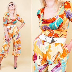 May include: A vintage jumpsuit featuring a vibrant floral pattern in orange, yellow, purple, and teal. The outfit includes a scoop-neck top with three-quarter sleeves, matching cropped pants, and a white belt with a silver buckle. The model is wearing sunglasses and heels.