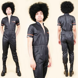 May include: Black jumpsuit with short sleeves, a mandarin collar, and a front zip. The jumpsuit has orange and red stripes on the sleeves and a small chest pocket. The model is wearing black boots.
