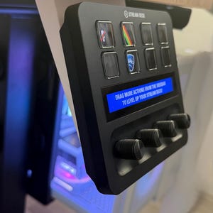Puede incluir: Panel de control Stream Deck negro con múltiples botones y perillas. El panel tiene una pequeña pantalla que muestra el texto "DRAG MORE ACTIONS FROM THE SIDEBAR TO LEVEL UP YOUR STREAM DECK!". Algunos botones tienen iconos. El dispositivo está montado en una superficie blanca.