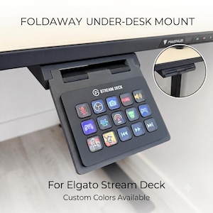May include: A black foldaway under-desk mount for an Elgato Stream Deck, featuring a hinged bracket that attaches to a desk edge. The device has fifteen square keys with various illuminated icons, including media controls and gaming symbols.