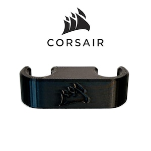 May include: A black 3D printed cable management clip featuring the Corsair logo embossed on the front surface. The object has a rectangular base with two circular cutouts on the top side, designed to hold cables securely in place for organization.
