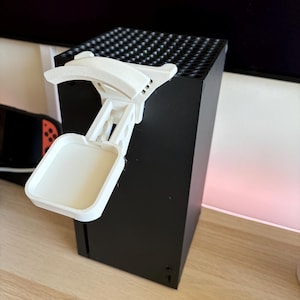 以下が含まれることがあります： 黒い長方形のゲーム機の上部に取り付けられた白いプラスチック製のヘッドセットスタンド。ヘッドセットを置くための湾曲した上部パーツと、本体前面から外側に突き出た四角いベースエクステンションを備えています。