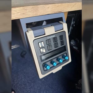 May include: A Stream Deck device with a beige and black frame, mounted under a wooden surface. The device has multiple buttons, a display, and blue knobs. The Stream Deck logo is visible.