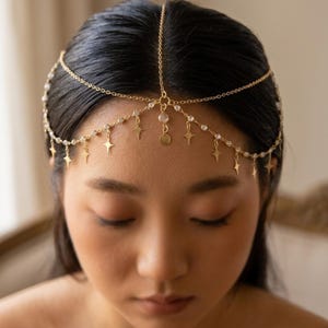 Può includere: Copricapo dorato con un delicato design a catena. Il copricapo presenta piccoli ciondoli a forma di stella, perline trasparenti e un pendente centrale. Il design è indossato sulla fronte e sopra la testa, creando un look elegante e celeste.