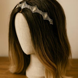 May include: Silver-toned metal headband with an ornate design featuring oval-shaped accents. The headband is displayed on a mannequin head with dark brown hair that fades to a lighter brown at the ends. The headband has a vintage or antique aesthetic.