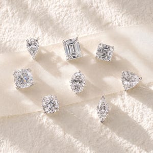 May include: A collection of eight clear, faceted gemstones in various cuts, including pear, emerald, square, round, oval, triangle, and marquise shapes, all set in silver-toned metal prongs and arranged on a smooth, light-coloured fabric ribbon.