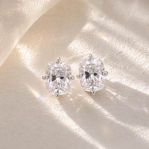 May include: A pair of silver-toned stud earrings featuring cushion-cut clear stones. Each earring is secured by four prongs, showcasing a faceted surface that reflects light. The design is symmetrical, with a polished metallic setting holding the stones.