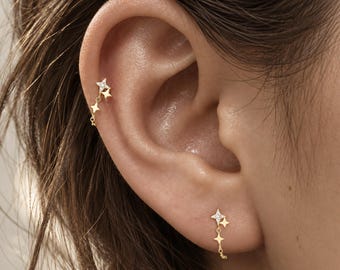 May include: A gold earring set featuring a star-shaped stud with a small clear stone, connected by a delicate chain to two additional dangling star charms. The design is worn in both the earlobe and the upper cartilage piercing of the ear.