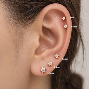 May include: Five gold flower-shaped stud earrings with clear stones, arranged in a row. The sizes are labelled as 2.8mm, 3.5mm, 3.5mm, 4.5mm, and 5.5mm. Each earring features a central gold bead surrounded by five small clear stones in a floral pattern.