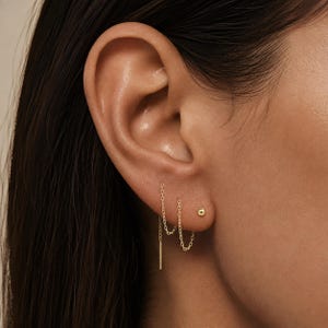 May include: A set of three gold earrings worn in one ear. The set includes a gold ball stud earring, a gold chain threader earring with a small dangling chain loop, and a gold chain threader earring featuring a thin, straight gold bar pendant.