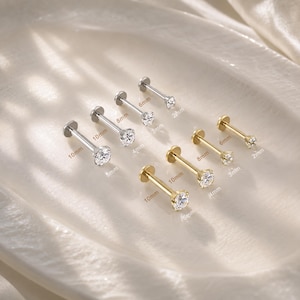 Könnte beinhalten: Eine Sammlung von silber- und goldfarbenen Labret-Steckern mit klaren, rund geschliffenen Edelsteinen. Jeder Stecker ist mit seiner spezifischen Länge von 6mm, 8mm oder 10mm und einem Steindurchmesser von 2mm, 3mm, 4mm oder 5mm beschriftet.