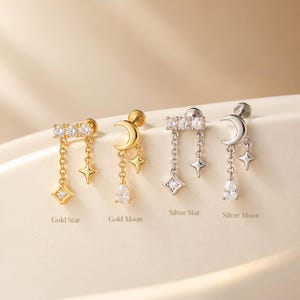 Op de afbeelding: Vier oorknopjes met platte achterkant, gelabeld Gold Star, Gold Moon, Silver Star en Silver Moon. De goud- en zilverkleurige oorbellen hebben bungelende kettinkjes met ster-, maan- en peervormige kristallen bedels en kleine steentjes.