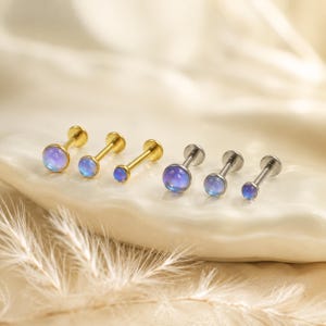 Könnte beinhalten: Sechs Labret-Piercings in zwei Dreierreihen mit runden, irisierenden Cabochon-Steinen. Die linke Gruppe besteht aus drei goldfarbenen Metallstäben, während die rechte Gruppe drei silberfarbene Metallstäbe mit flacher Rückseite umfasst.