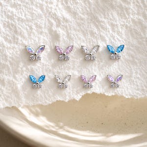 Puede incluir: Ocho pequeños pendientes de botón con forma de mariposa dispuestos en dos filas. Cada pendiente presenta dos piedras preciosas de talla marquesa para las alas y dos piedras redondas transparentes para el cuerpo, sobre una base de metal plateado.