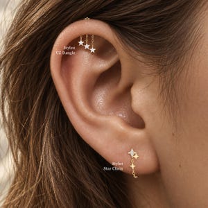 Op de afbeelding: Goudkleurige oorsieraden in twee verschillende ontwerpen: Style 2 CZ Dangle, een helix-piercing met drie bungelende sterbedels bezet met heldere stenen, en Style 1 Star Chain, een oorbel met twee verbonden sterbedels aan een fijn kettinkje.
