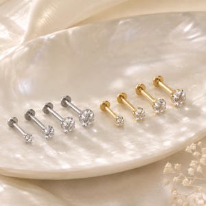 May include: A set of eight flat-back stud earrings arranged in two rows. Four earrings are silver-toned and four are gold-toned, each featuring a single clear round-cut gemstone set in a prong setting at the end of a straight metal post.