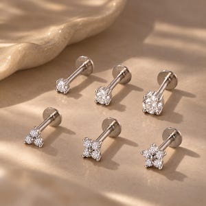 Op de afbeelding: Een verzameling van zes zilverkleurige labret-piercings met platte, ronde achterkantjes en decoratieve voorkanten. Drie piercings hebben een enkele ronde, heldere steen in verschillende maten, terwijl de andere drie clusters van steentjes in driehoekige, vierkante en bloemvormen bevatten.