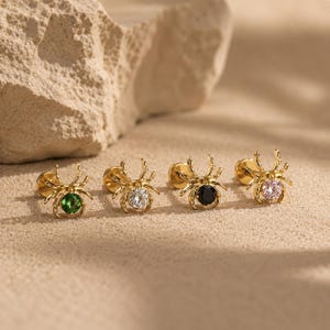 May include: Four gold-toned spider-shaped stud earrings with flat back posts. Each spider features a round faceted gemstone in the center, specifically one green, one clear, one black, and one light pink stone, arranged in a horizontal row.