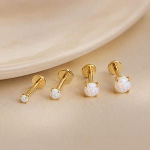 May include: Four gold-tone flat back labret studs arranged in a row, each featuring a round, iridescent white opal gemstone set in a four-prong setting. The studs vary in size, with the smallest on the left and the largest on the right.