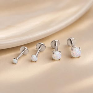 May include: Four silver-toned labret studs arranged in a row, each featuring a round, iridescent white opal-like stone held by four prongs. The studs have a flat circular back and a straight post, with the stones increasing in size from left to right.