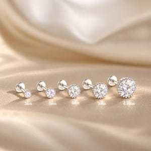 Pode incluir: Cinco brincos de prata com pedras redondas transparentes, cada um com um fecho plano marcado com S925. As pedras estão cravadas numa moldura de contas e variam em tamanhos de 2mm, 3mm, 4mm, 5mm e 6,5mm, conforme indicado abaixo de cada um.