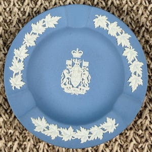 May include: A round, light blue ashtray with a white leaf border and a white emblem in the center. The emblem features a crown and a coat of arms. The ashtray is made of ceramic.