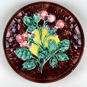 May include: A round ceramic plate featuring a dark brown basket-weave texture on the rim. The center displays a raised, colorful relief of green leaves, small pink flowers, and a cluster of yellow berries, all finished with a glossy glaze.