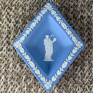 May include: A diamond-shaped, light blue trinket dish with a white floral border and a white classical figure in the center. The dish is made of ceramic or porcelain and is likely vintage.