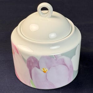 May include: A white ceramic sugar bowl with a lid and a loop handle. The bowl is decorated with a floral pattern of pink and purple flowers on a gray background. The sugar bowl is cylindrical in shape.