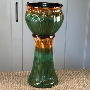 May include: A green ceramic planter on a pedestal. The planter has a glossy finish with brown and black accents. The pedestal is a matching green with brown and tan stripes. The planter is designed for indoor or outdoor use.