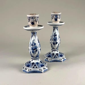 May include: A pair of ceramic candlesticks featuring a white base with intricate blue floral and geometric patterns. Each candlestick has a flared base, a bulbous central stem, and a circular top cup designed to hold a single taper candle.