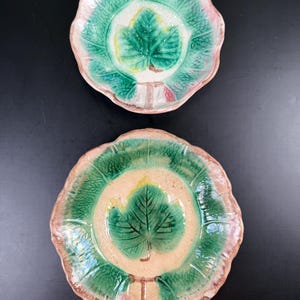 May include: Two ceramic leaf-shaped plates. The plates feature a central green leaf design with a cream-colored background, surrounded by a green, textured border. The rims are edged with pink and brown tones. The plates are on a black surface.