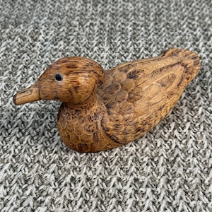 May include: A carved wooden duck figurine with detailed feather patterns and a small black eye. The duck is a warm brown color and has a smooth, polished finish. The duck is a decorative item.