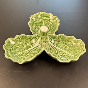 May include: A green ceramic serving dish in the shape of a cabbage. The dish has three leaf-shaped sections radiating from a central stem. The leaves have detailed veining and a glossy finish.