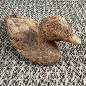 May include: A detailed wooden duck figurine, featuring carved feather details and a natural brown finish. The duck is positioned with its head turned, showcasing its beak and textured head. A decorative item for home decor.