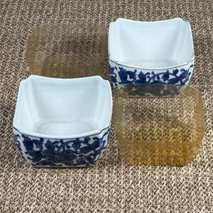 May include: Four square ceramic bowls with blue floral designs on a white background. Two clear, square lids are also present. The bowls have a slightly curved rim and are likely for serving or display.