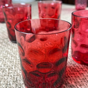 May include: A set of six red drinking glasses. Each glass has a textured, patterned design. The glasses are made of a transparent red material, allowing light to pass through. The glasses are cylindrical and appear to be of a medium size.