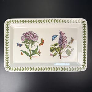 May include: Rectangular serving tray with a white background, featuring botanical illustrations of flowers and butterflies. The tray has a green leaf border and includes the scientific names of the plants. A decorative piece for serving or display.