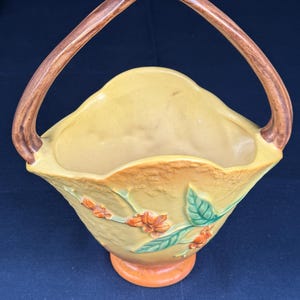 May include: A yellow ceramic basket with a brown handle. The basket features embossed orange flowers and green leaves on a branch. The base is orange. The handle is arched and made of wood.