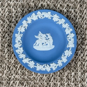 May include: A small, round blue plate with a white relief design. The plate features a central scene of figures and a border of white floral and leaf patterns. The plate is likely made of ceramic or porcelain.