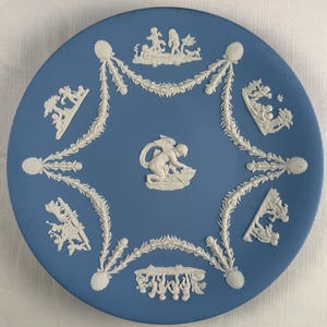 May include: A blue ceramic plate with white relief decorations. The plate features a central cherub figure and surrounding garland designs. Additional white relief scenes are placed around the plate's edge, creating a decorative border. The plate has a smooth, matte finish.