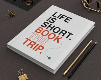 May include: A white hardcover journal featuring the black text "LIFE IS SHORT." and the orange text "BOOK THE TRIP." with a dotted line leading to a small teal airplane icon. The journal is accompanied by gold binder clips and two pencils.
