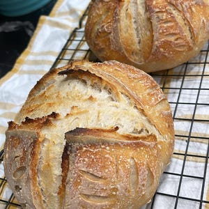 Pode incluir: Duas broas de pão artesanal recém-assadas, douradas com a parte superior marcada, repousam sobre uma grelha de arame preta sobre um pano xadrez amarelo e branco. O pão tem um aspeto rústico, com uma crosta crocante e um interior macio.
