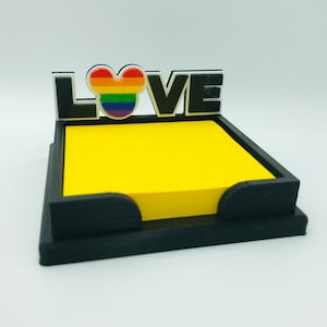 May include: Black memo pad holder with the word "LOVE" in black and white lettering. The "O" is a rainbow-colored Mickey Mouse head. The holder contains a stack of yellow sticky notes.