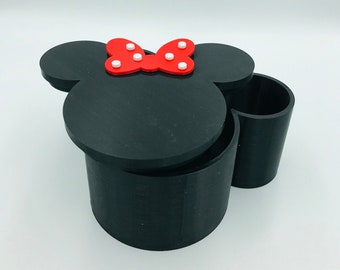Könnte beinhalten: Schwarzer und roter Aufbewahrungsbehälter im Minnie-Mouse-Design mit abnehmbarem Deckel. Der Behälter hat eine runde Form mit zwei Ohren und einer Schleife oben.