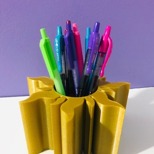 May include: A gold-colored, sun-shaped pen holder filled with various colorful pens. The pens include green, blue, purple, and pink, with the text "Pilot G-2 07" visible on some. The holder sits on a white surface against a purple background.