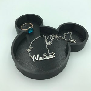 Pu&ograve; includere: Vassoio portagioie nero a forma di Topolino con un anello e una collana. L'anello ha una fascia dorata e una pietra preziosa blu. La collana d'argento riporta il nome "Melissa". Il vassoio &egrave; realizzato in materiale scuro.