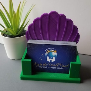 May include: Green and purple 3D printed business card holder shaped like a seashell. The holder has a slot for business cards and a decorative top. The business card holder has the text "Key to the World Travel" and "your key to a magical vacation".